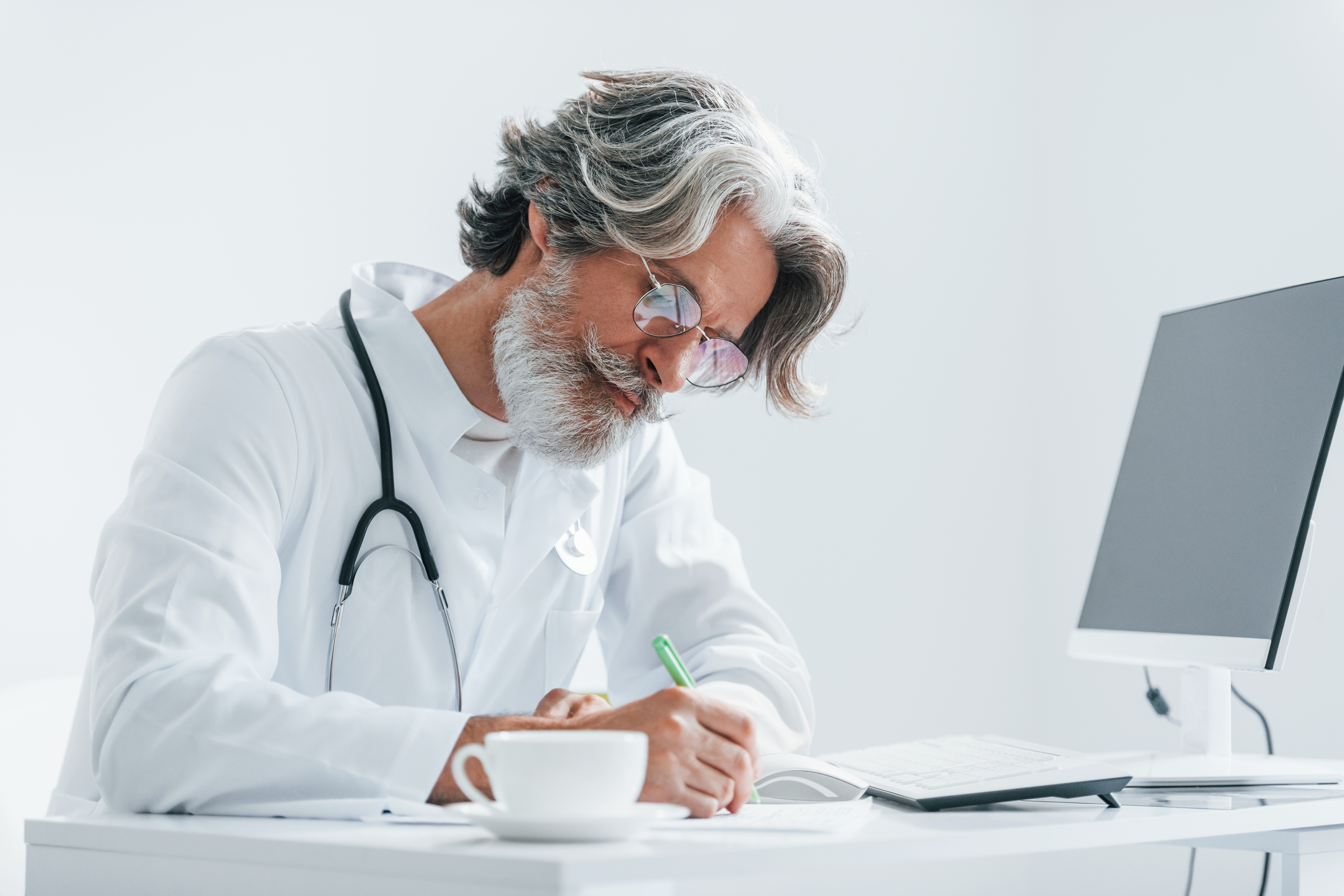 Escribiendo en la mesa del ordenador. Médico mayor con cabello y barba canosos, en bata blanca, dentro de la clínica.